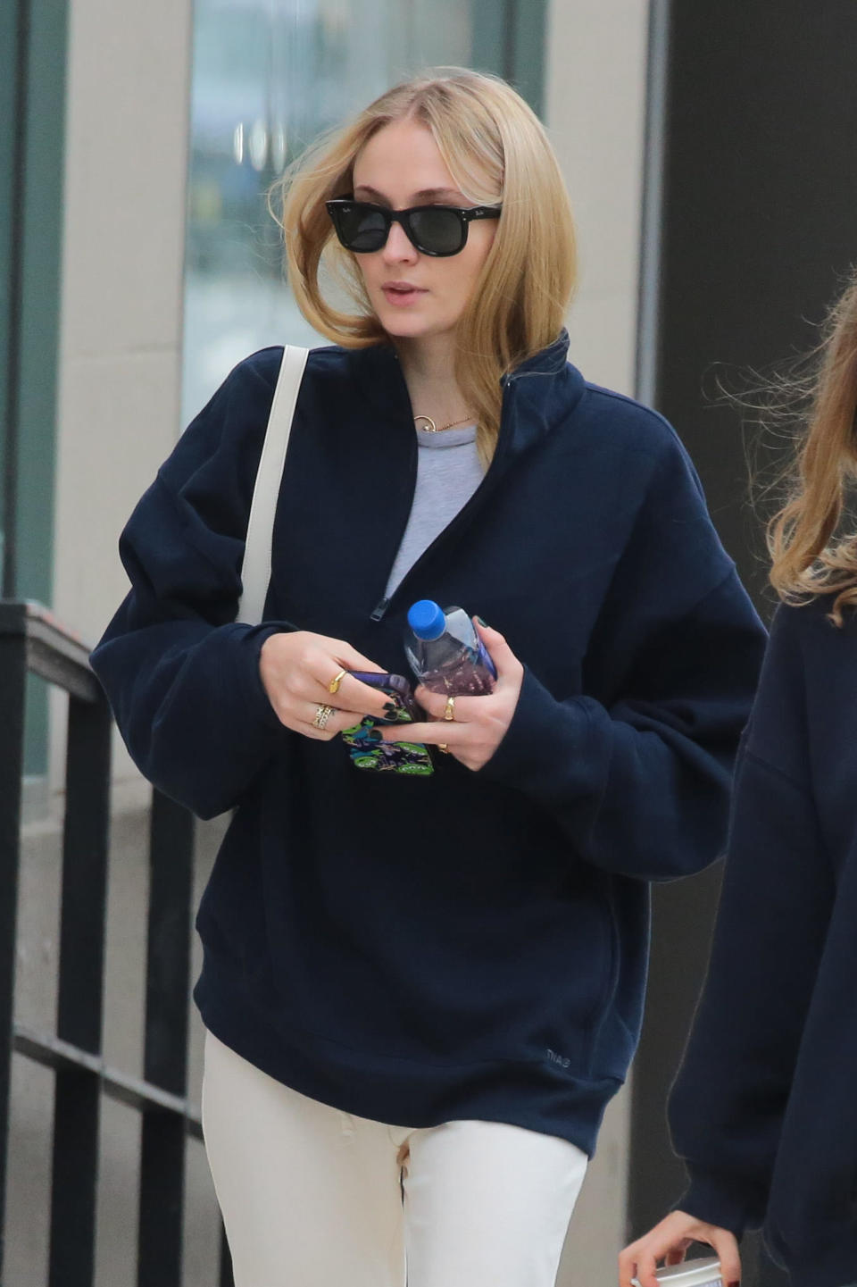 Sophie Turner walks outside wearing a dark pullover, sunglasses, and holding a water bottle
