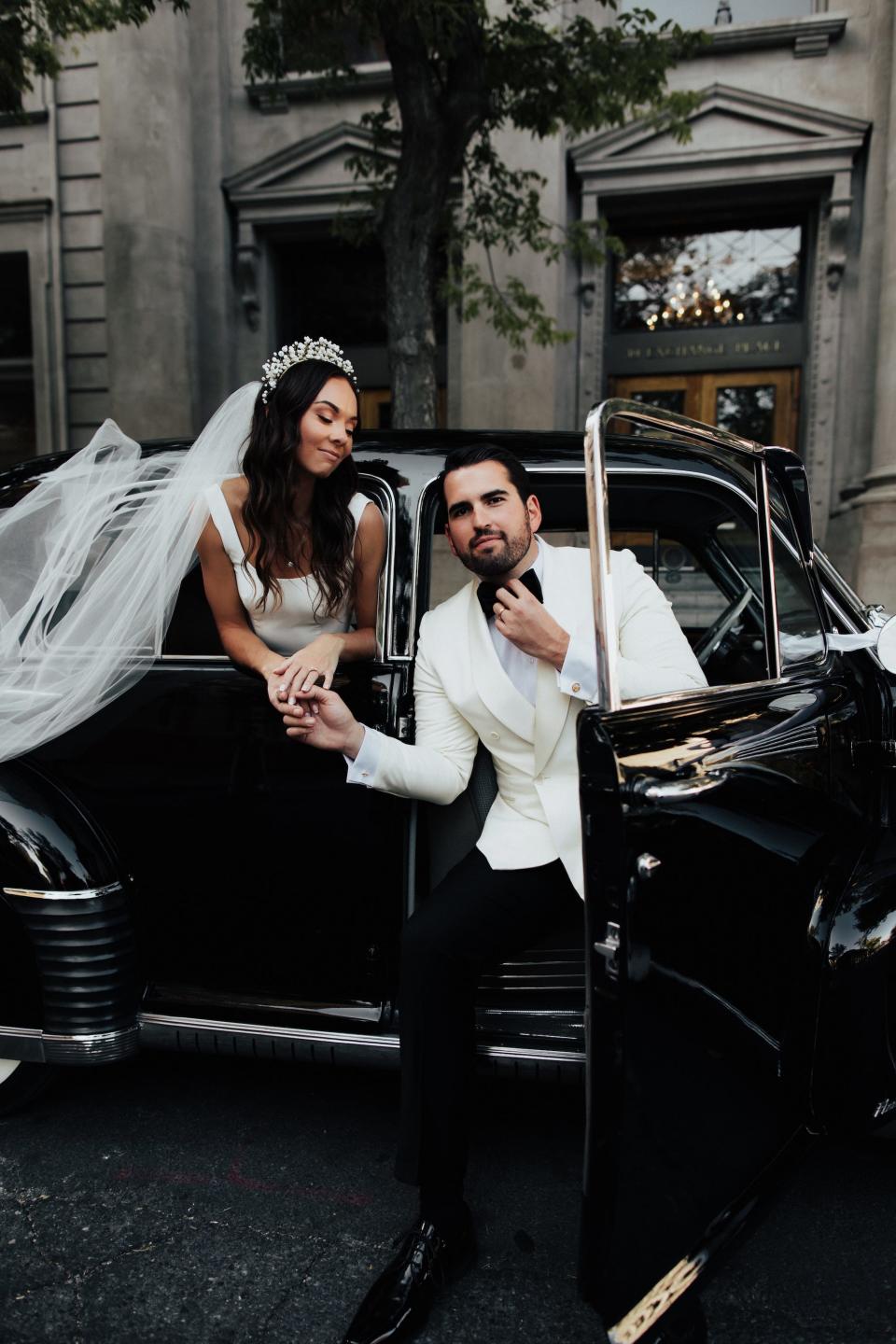 A bride and groom hold hands and pose in a car on their wedding day.