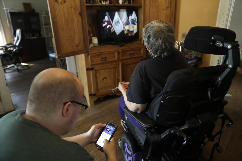 In this Tuesday, April 28, 2020, photo Zach Stafford, left, checks the news on his phone while sitting alongside his mother, Debra Mize, as they watch a livestream of the daily coronavirus briefing by Illinois Gov. J.B. Pritzker on the television in Belleville, Ill. The pair say they are consuming hours of news a day in various formats about the coronavirus. Americans are grappling with an essential question as they try to get the information they need to stay safe during the coronavirus crisis: Whom do you trust? (AP Photo/Jeff Roberson)