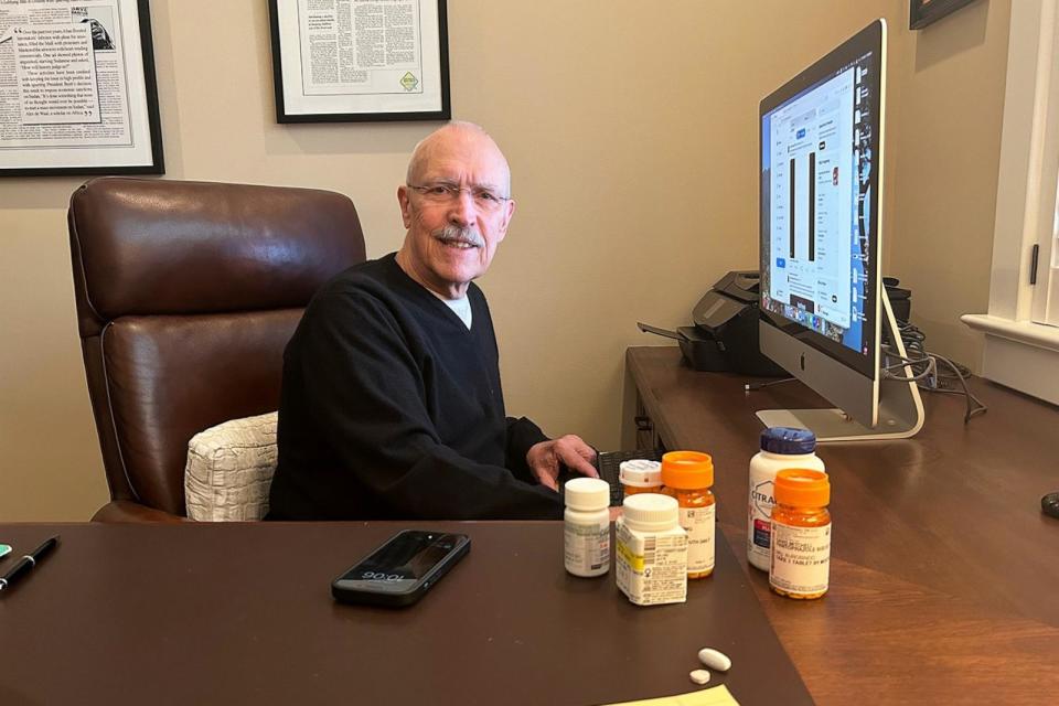 PHOTO: David Mitchell, founder of Patients for Affordable Drugs, sits in his home office. Beside him are some of the many drugs he takes to treat multiple myeloma and other conditions. (Arthur Allen/KFF Health News)