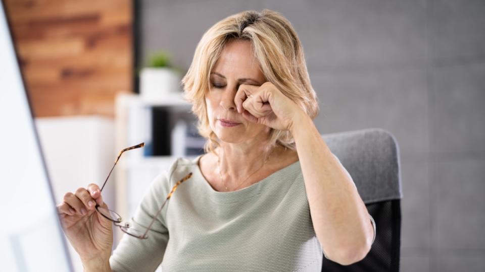 A woman with short blonde hair holding her glasses in one hand and rubbing her eye with the other