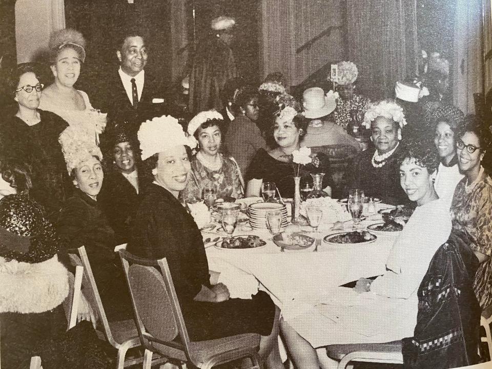 in a black and white photo, people in fancy outfits pose at a dinner table