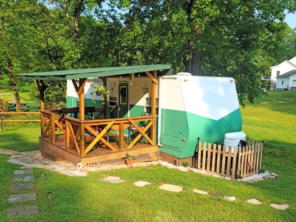 A green and white RV with a wooden porch. The porch has a green roof and the RV is located in a residential, grassy area with trees.