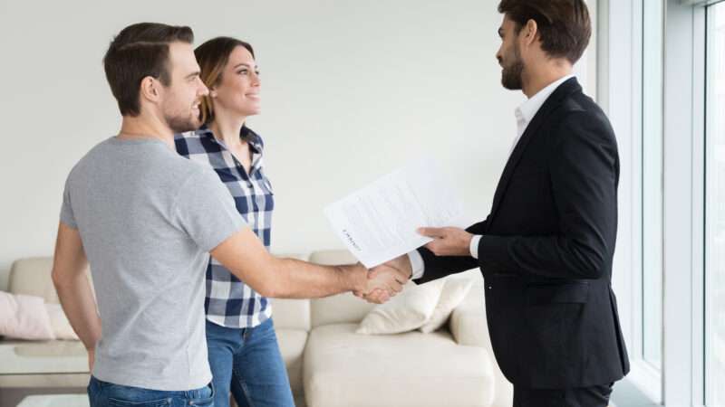 A realtor or landlord shakes hands with a young couple in a bright, sparsely-furnished apartment, while holding a rental agreement.