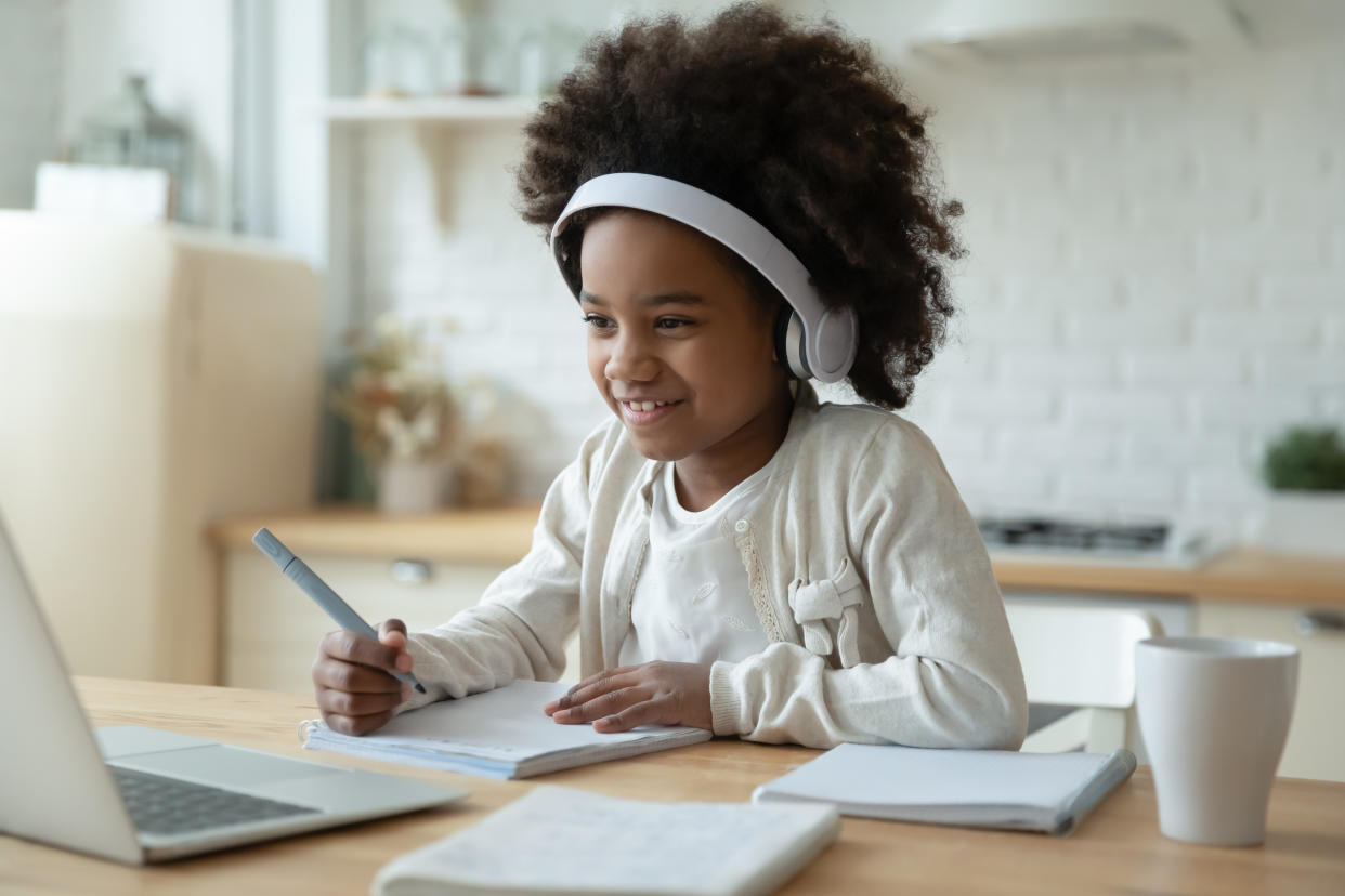 Smiling small African American girl in headphones watch video lesson on computer in kitchen, happy little biracial child in earphones have online web class using laptop at home, homeschooling concept