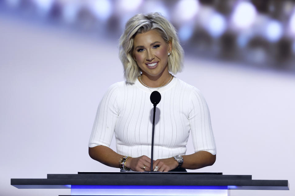 Savannah Chrisley steht in einem figurbetonten weißen Kleid hinter einem Podium mit einem Mikrofon und lächelt während einer Rede oder Präsentation