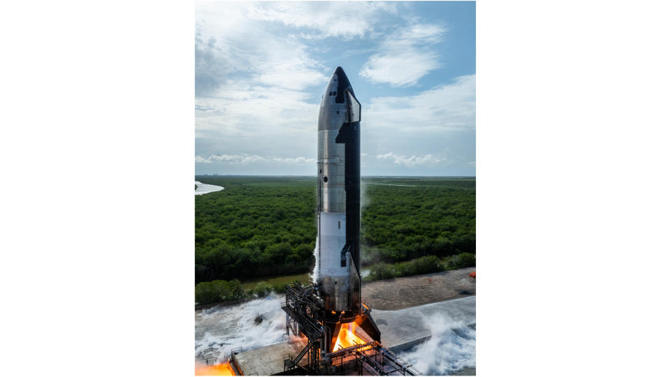 a large silver rocket fires its engines on an outdoor test stand, with greenery stretching into the distance