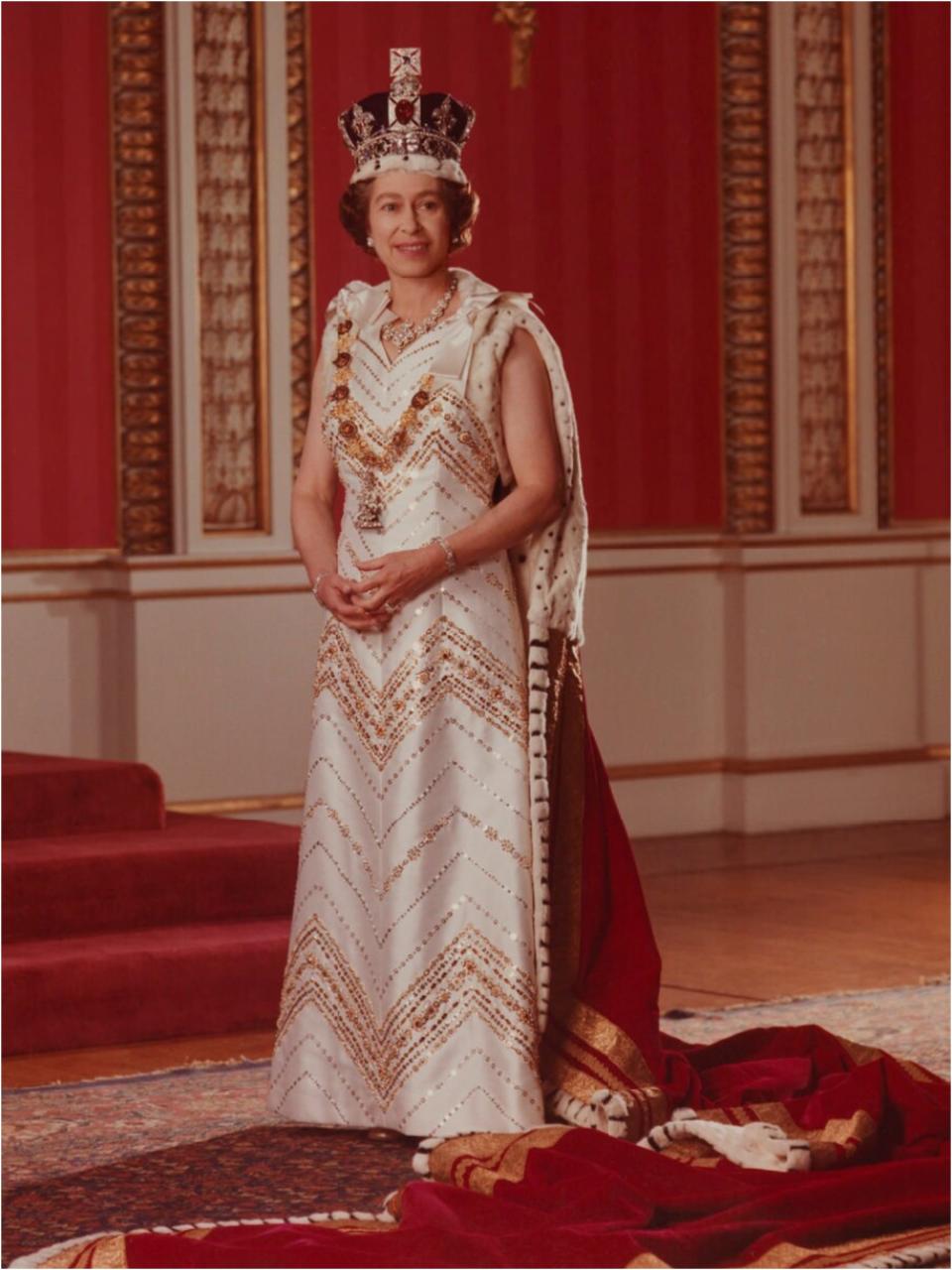 The Queen standing and smiling in an ornate crown and a white and gold sleeveless gown with a red long cape.