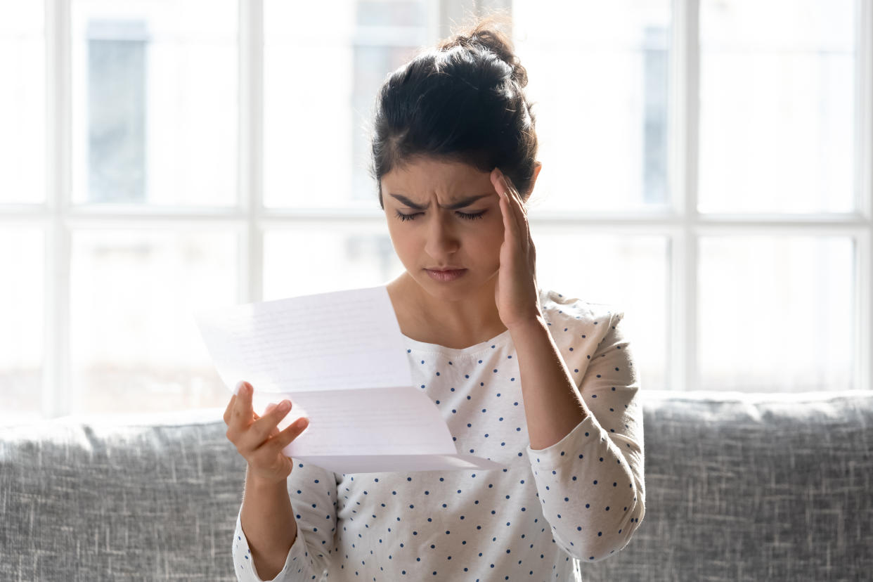 Stressed millennial indian woman reading paper letter with bad news, head shot close up. Unhappy young girl received money bank debt notice or eviction notification, feeling unwell, sitting on couch.