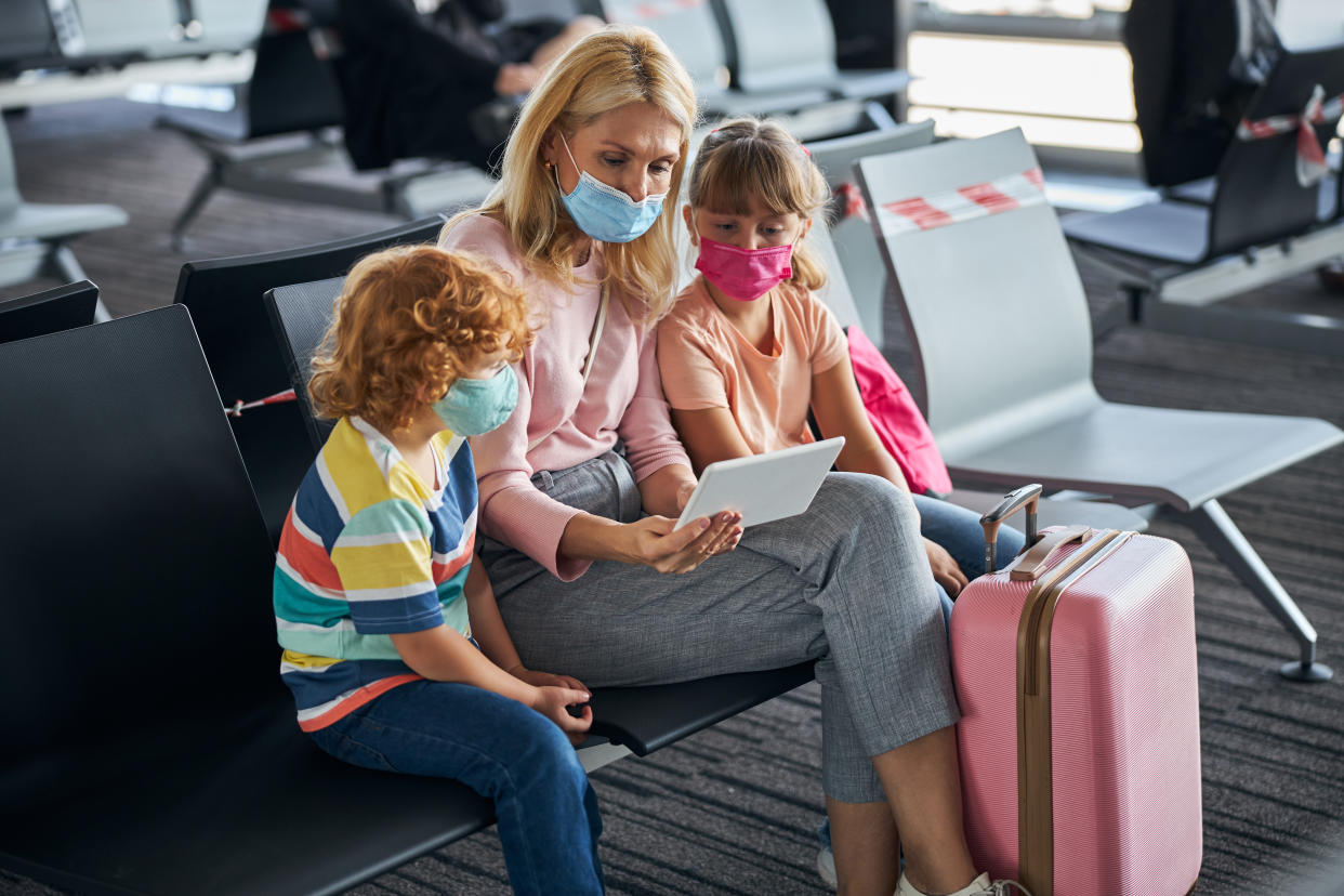 Woman and two children wearing medical face masks while sitting on plastic chairs and focusing on a tablet screen