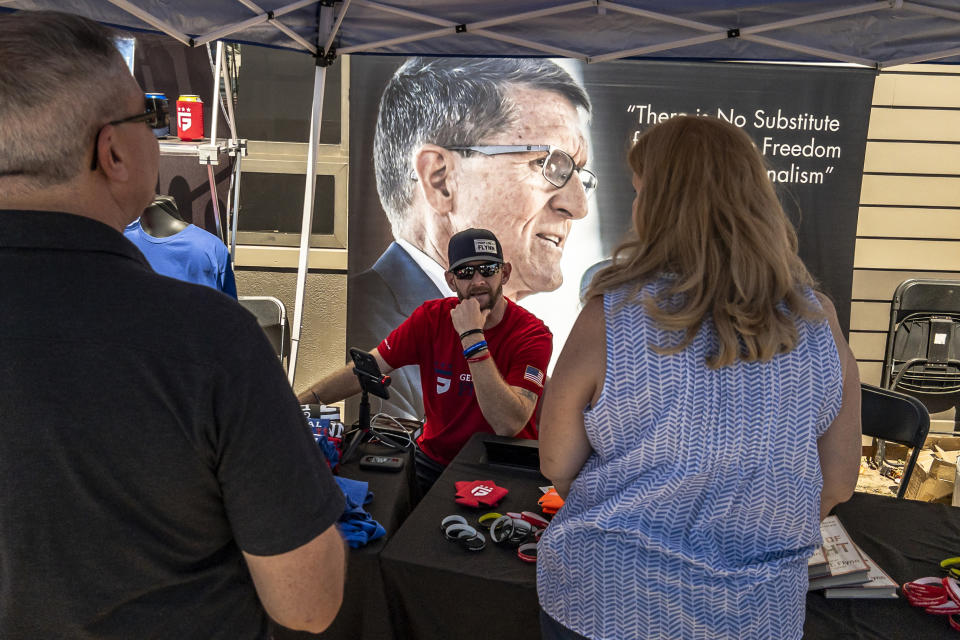 Michael Flynn, Jr., talks to people as he sits at the merchandise booth for his father, a retired three-star general who served as Trump's national security adviser, during the ReAwaken America tour at Cornerstone Church, in Batavia, N.Y., Saturday, Aug. 13, 2022. The elder Michael Flynn has drawn together election deniers, mask and vaccine opponents, insurrectionists, Proud Boys, and elected officials and leaders in state and local Republican parties. Along the way, AP and Frontline documented, Flynn and his companies have earned hundreds of thousands of dollars. (AP Photo/Carolyn Kaster)