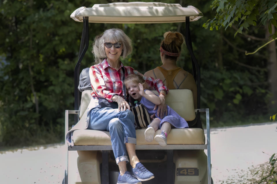 An older woman and a small child ride in the back of a golf cart, while a younger woman drives. The child, dressed in a dress, is resting on the older woman's lap