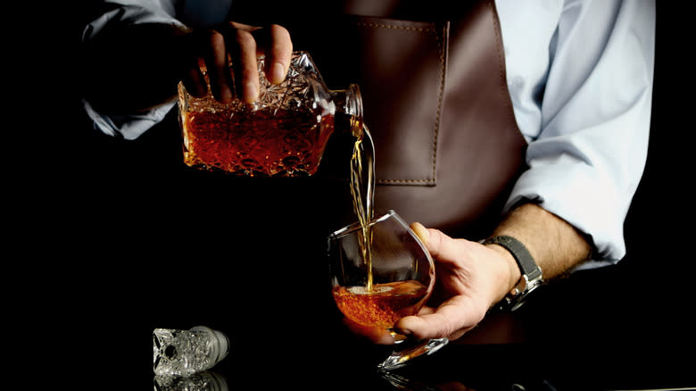 A bartender pouring brandy from a glass bottle into a snifter