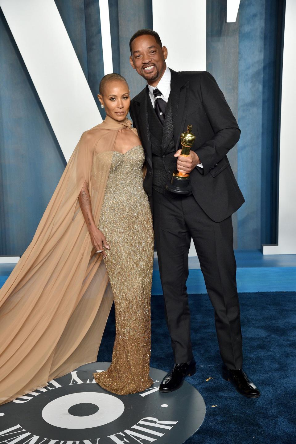 Jada in a gold ombre crystal sweetheart gown. She's wearing a brown see-through cape that ties around her neck and flows down to a long train. Will is wearing a black three-piece suit with a crustal band and pin on his black tie.