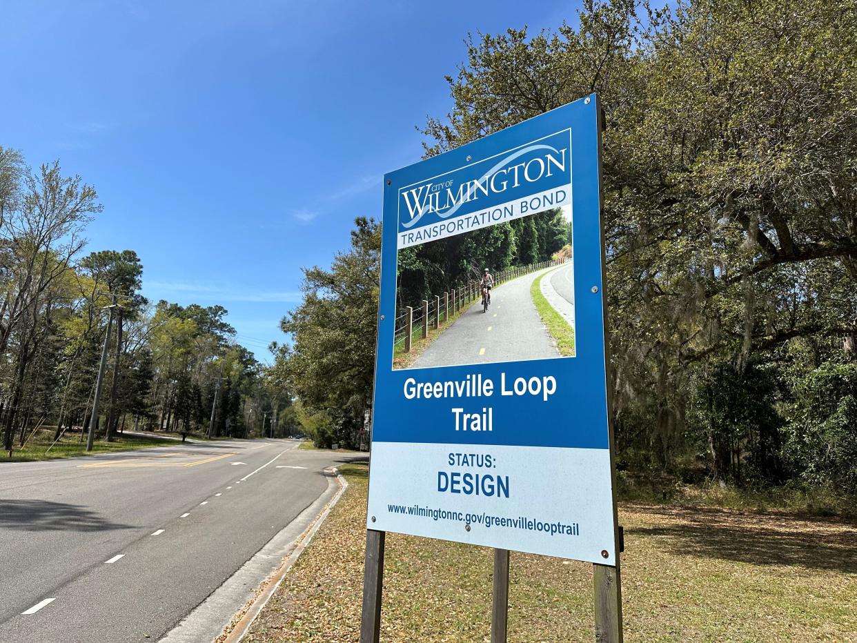 A Greenville Loop Trail status sign sits alongside Greenville Loop Road in front of Bradley Creek Elementary School. The sign lists the project status as in the design phase.