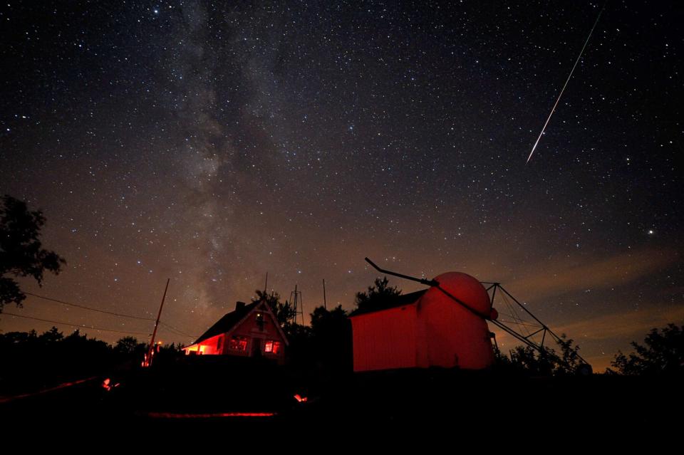 A meteor streaking across a dark-purple night sky above a house and a building illuminated in red.