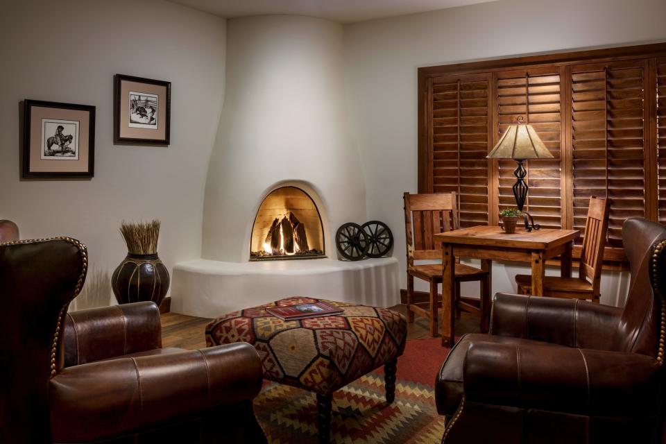 A room with brown leather chairs in front of a white, adobe, lit fireplace