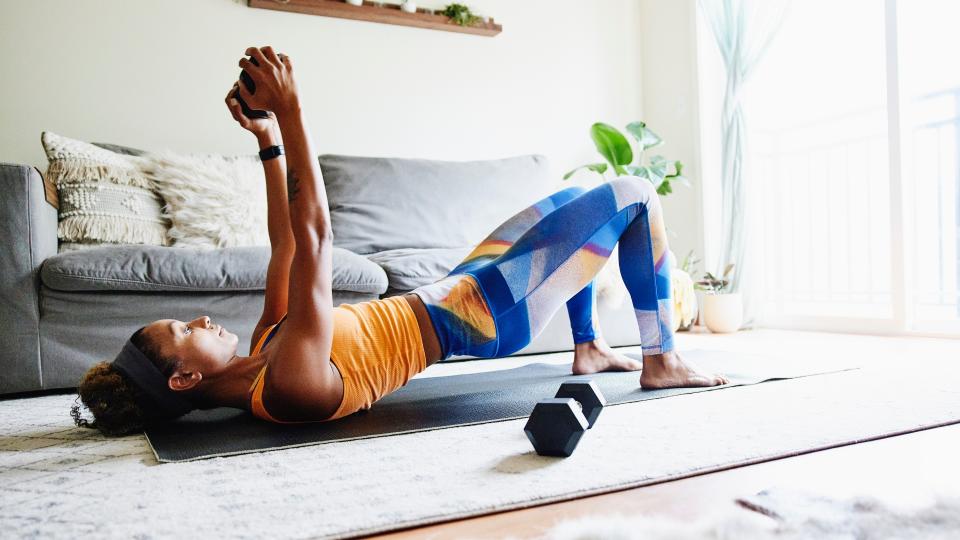 A woman lies on her back on a living room floor, one weight at her side and holding another in the air over her chest.