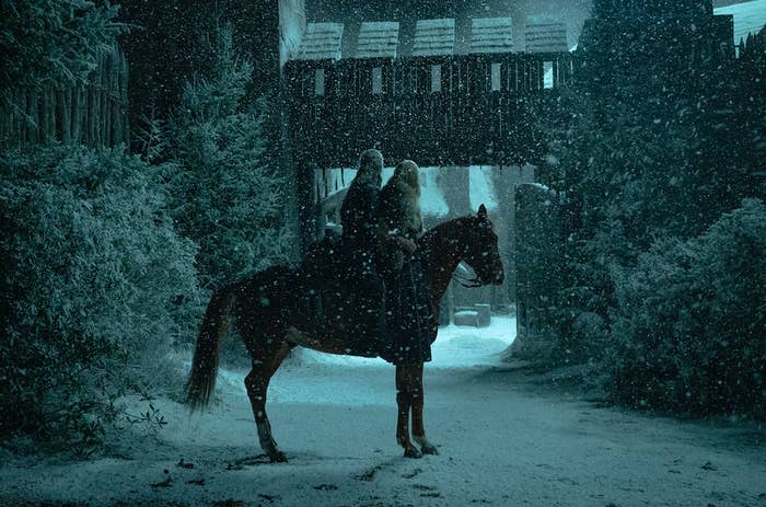 The Witcher on a horse with a companion looking up at a structure as it snows