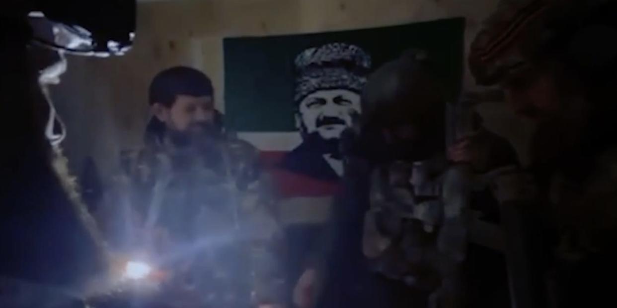 A still from video posted by Grozny TV showing Chechen leader Ramzan Kadyrov in a darkened room with soldiers. A flag printed with an image of his father hangs behind them.