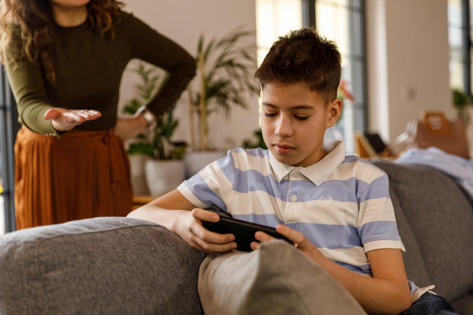 A boy focused on his handheld game with an adult standing beside him, gesturing as if speaking