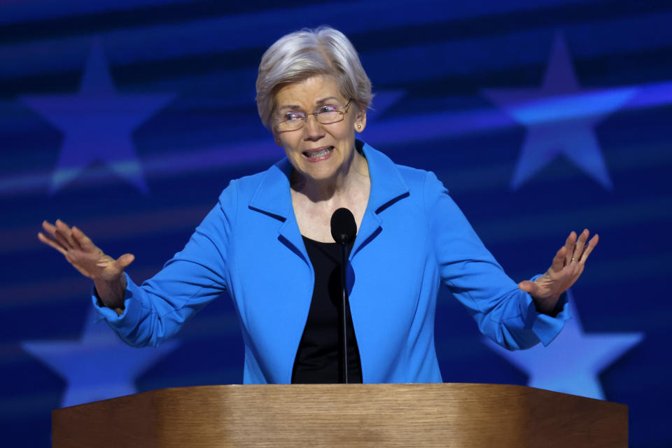 Elizabeth Warren speaks at a podium with her hands slightly raised. She is wearing a blazer