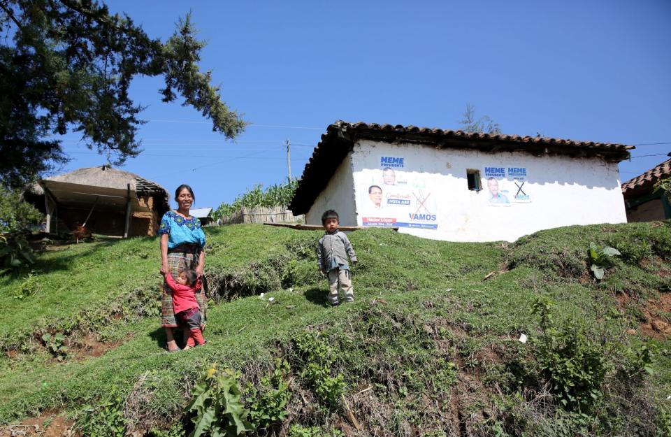 A woman and two children stand outside a mud brick home with political signs on the wall.