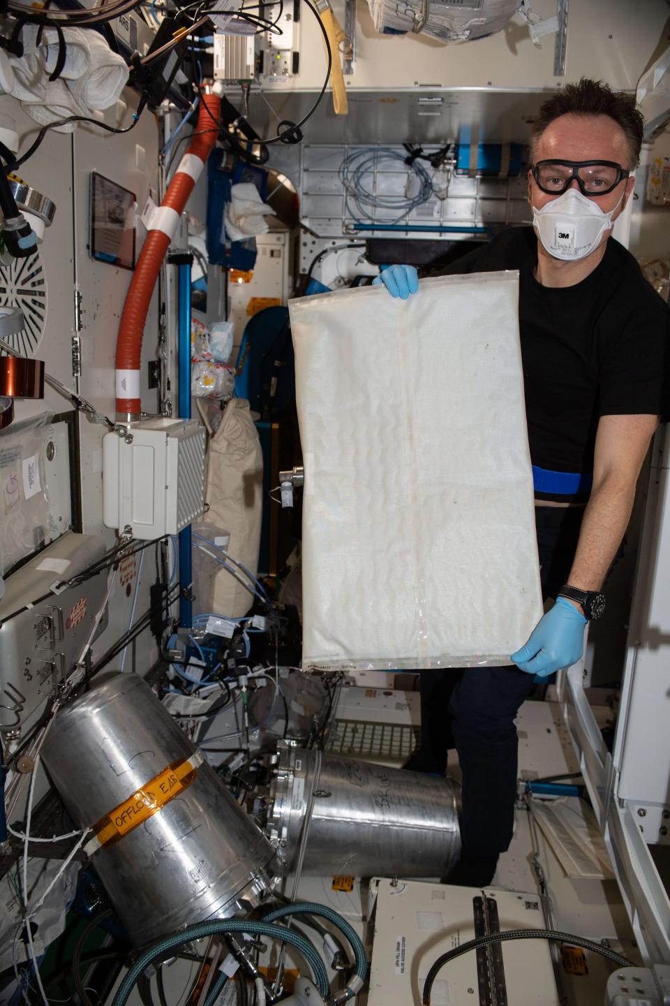 an astronaut in n95 and goggles and medical gloves holding a large cotton-tipped rectangle in a small room inside the space station