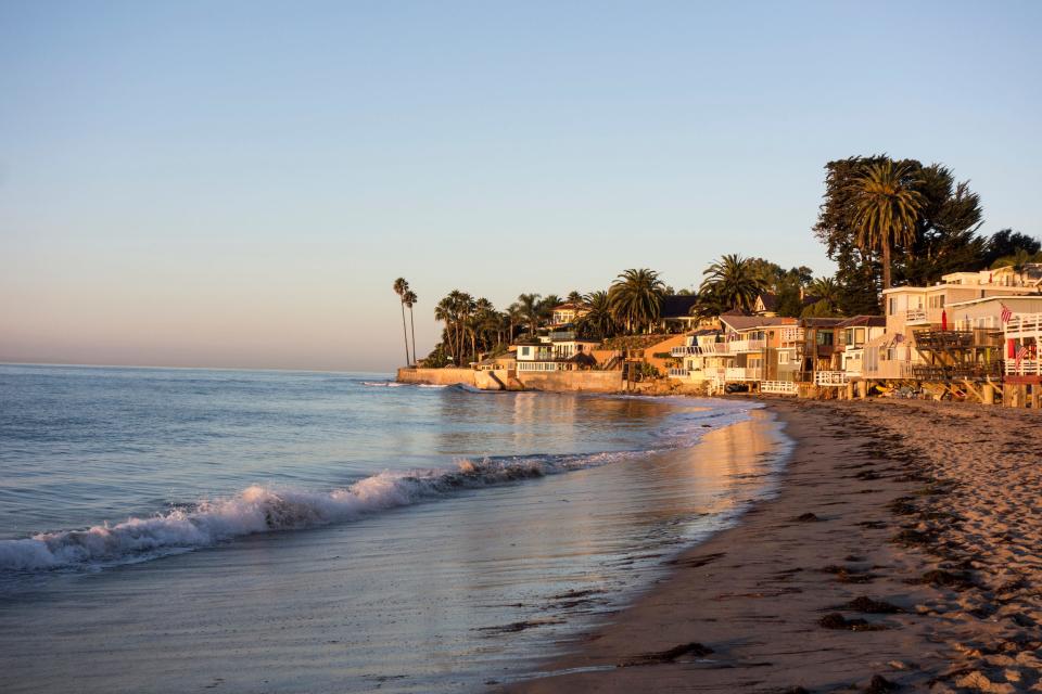 An empty beach with gentle waves and houses and palm trees along the coast