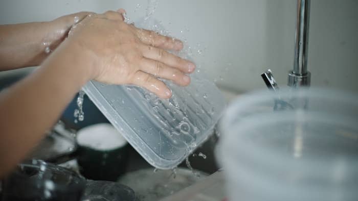 Person washing a plastic container under running water in a kitchen sink, with splashes visible