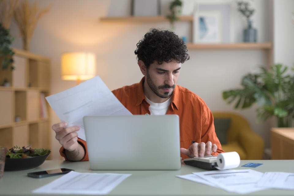 A person in a red shirt works at a desk with a laptop, holding papers and using a calculator in a home office setting