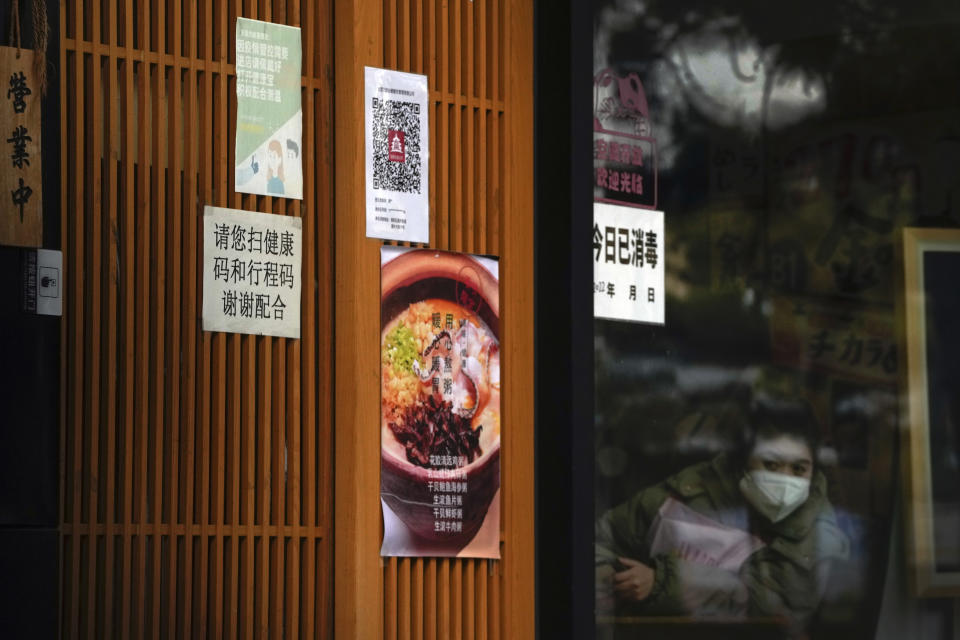 A masked worker looks out from a restaurant as its entrance door displays a health check QR code and a notice that reads "Please scan your health QR code and travel code, thank you for your cooperation" in Beijing, Monday, Dec. 12, 2022. China will drop a travel tracing requirement as part of an uncertain exit from its strict "zero-COVID" policies that have elicited widespread dissatisfaction. (AP Photo/Andy Wong)