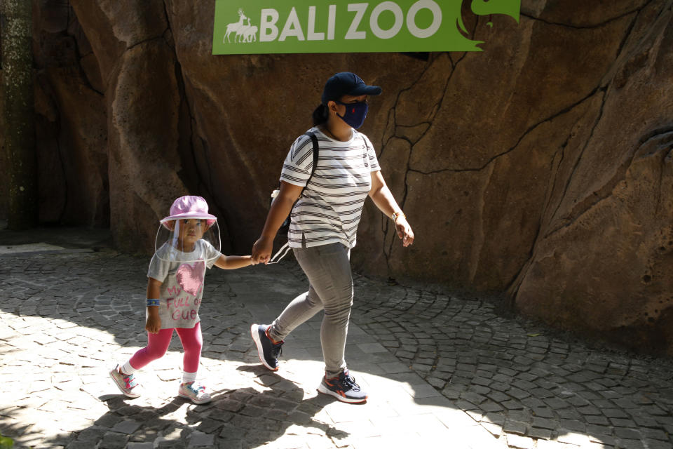 A mother walks with her daughter wearing protective gear during their visit to the zoo in Bali, Indonesia, Monday, July 13, 2020. Indonesia's resort island of Bali reopened after a three-month virus lockdown last week, allowing local people and stranded foreign tourists to resume public activities before foreign arrivals resume in September. (AP Photo/Firdia Lisnawati)