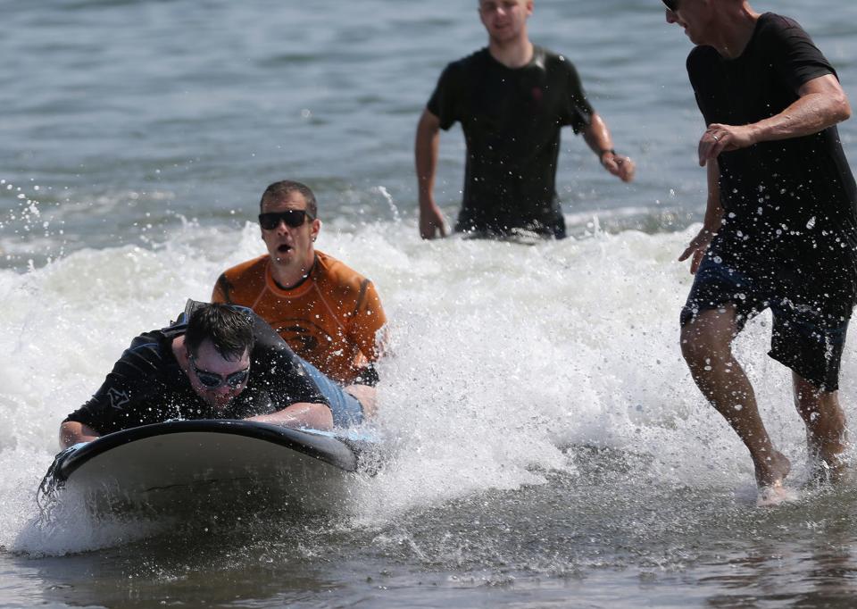 Veteran Mico Morin holds tight to the surf board as volunteers help guide  him during the Wounded Warrior Project Hit The Beach event, which helps injured veterans learn how to surf at North Beach in Hampton Aug. 27, 2021.