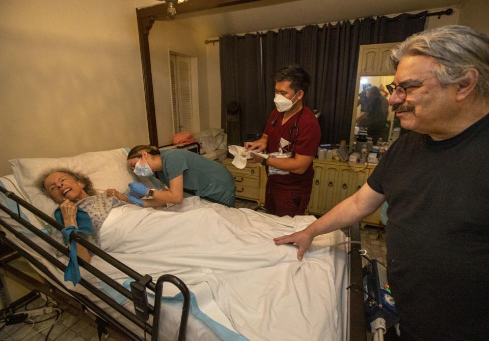 A bedridden woman gets a shot while her son touches her foot.