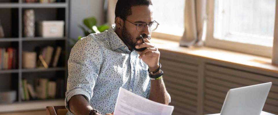 Focused African-American man holding paper and looking at laptop computer