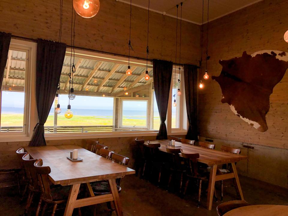 An interior of a restaurant with two tables, each with 6 chairs. Lightbulbs hang from the ceiling, cow skin is hung on the wall, and a large window offers a picturesque view.