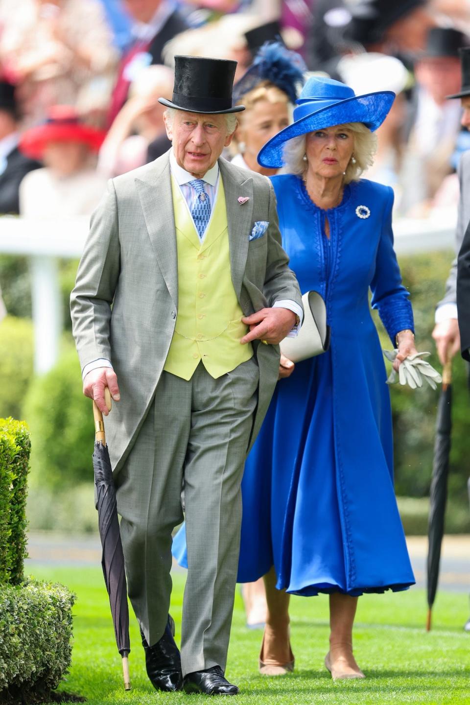 King Charles III, wearing a gray morning suit with a yellow waistcoat and a black top hat, walks beside Queen Camilla, wearing a royal blue long-sleeved dress with a coordinating hat.