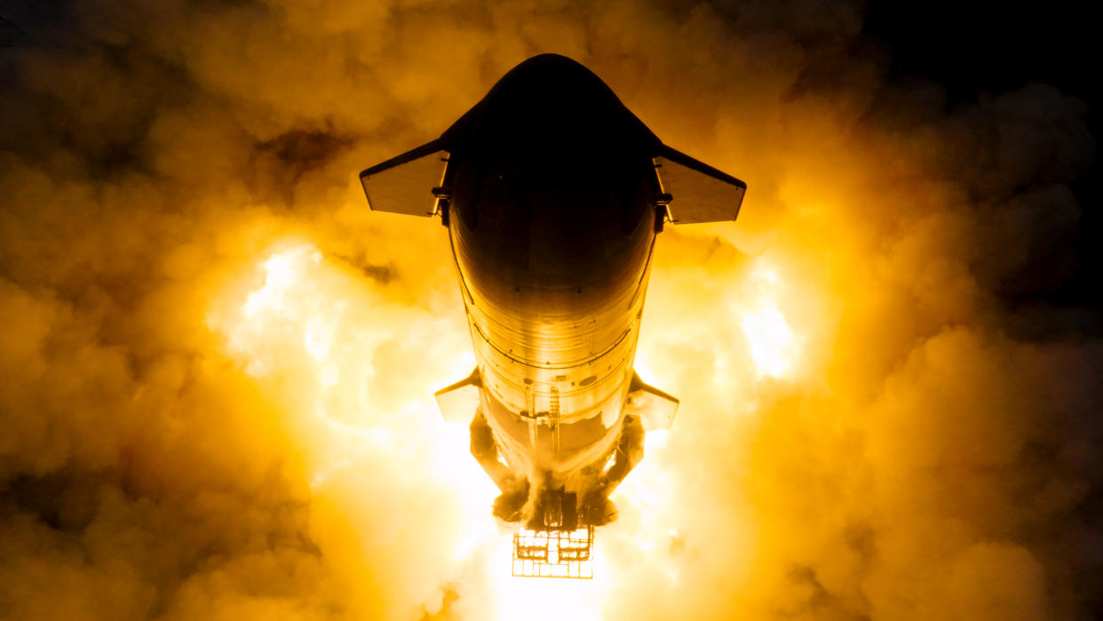  Photo looking down on a cylindrical silver rocket with orange flames erupting from its base. 