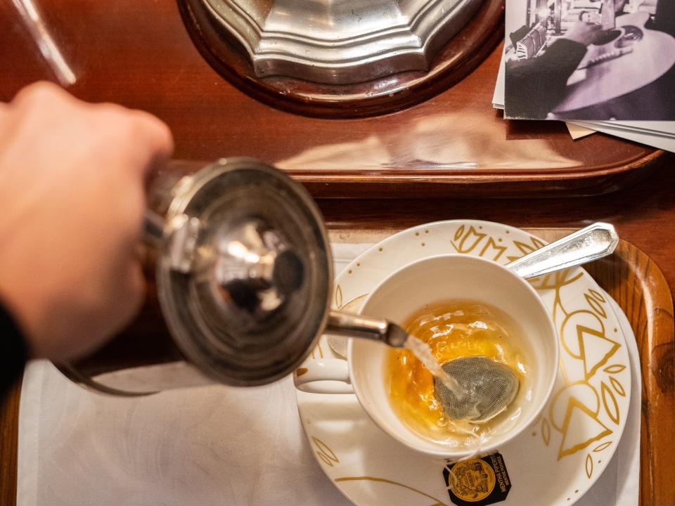 An aerial view of a hand pouring a silver pot of hot water into a teacup on a dark, brown wooden table.