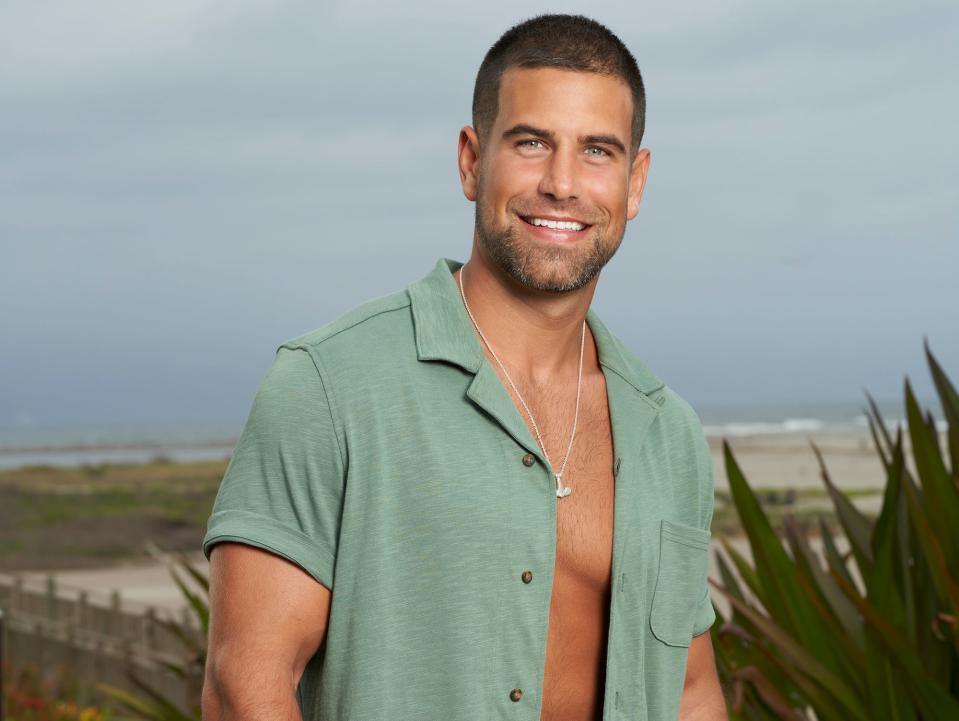 Blake Moynes poses in a green unbuttoned shirt and black shorts.