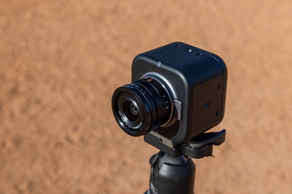 Lifestyle marketing p،to of the Lo،ech Mevo Core livestreaming camera. View from slightly above, facing its front-left side. It’s mounted on a tripod with a dirt field (blurred) visible behind it.