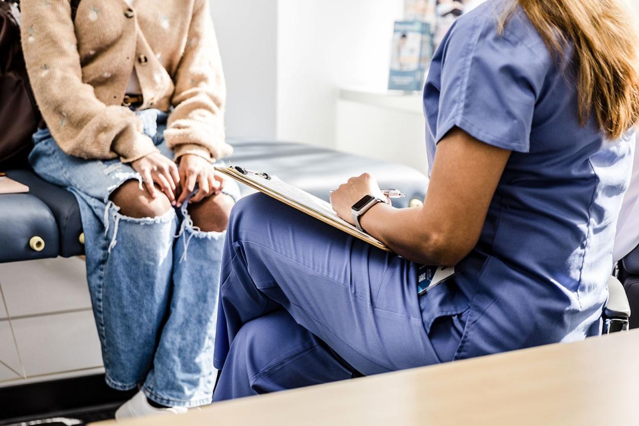 A female doctor in scrubs sits with her legs crossed and hands across a medical pad as a woman in torn jeans and a button-up sweater sits on a patient's table.