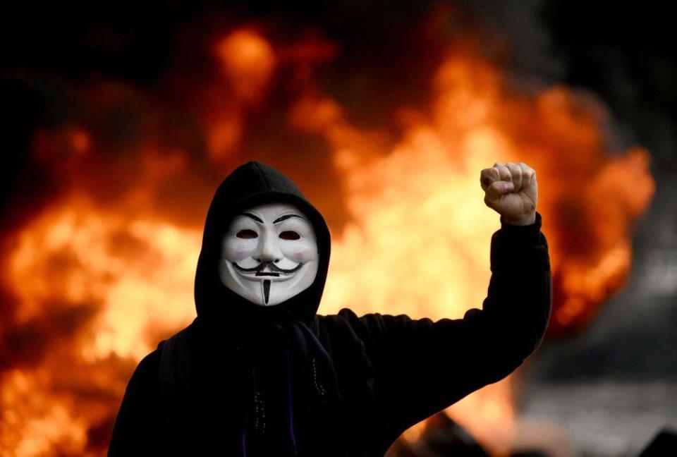 Una persona con la careta de Anonymous y un incendio detrás