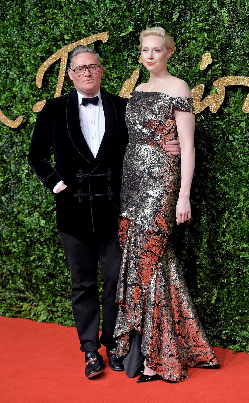 Gwendoline Christie in a metallic floral gown and heels standing taller than Giles Deacon in a velvet tuxedo.