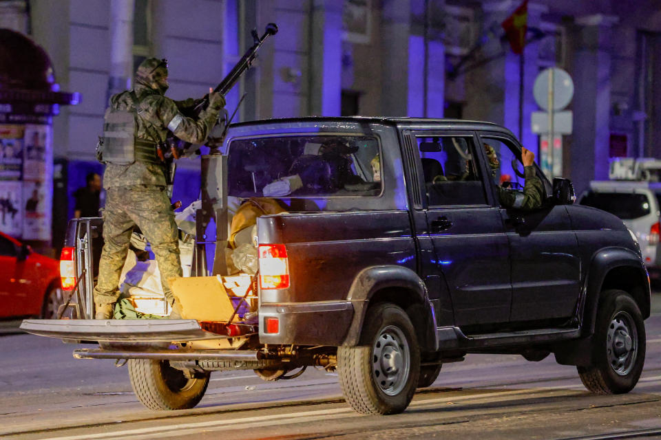 Someone in military gear stands on the back of a pickup truck holding a weapon.