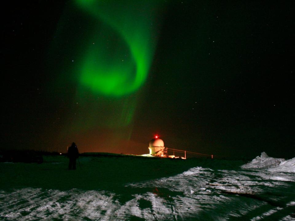 green spiral ribbon aurora borealis in the night sky above a snowy field with a person and a small white observatory