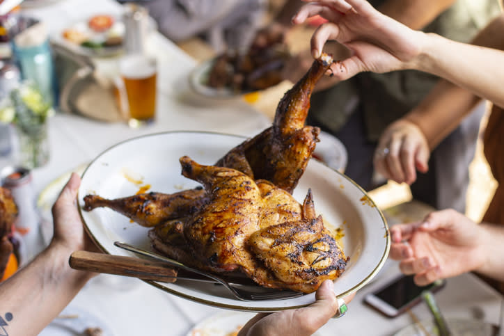 Grilled, whole, barbecued chicken on a platter being served at a table with hands reaching in, beer glasses and other dishes in the background