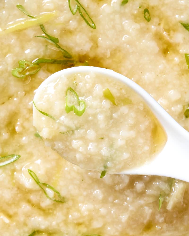 Overhead shot of congee in a white bowl, topped with green onion and theres a white soup spoon resting on the right side of the bowl.