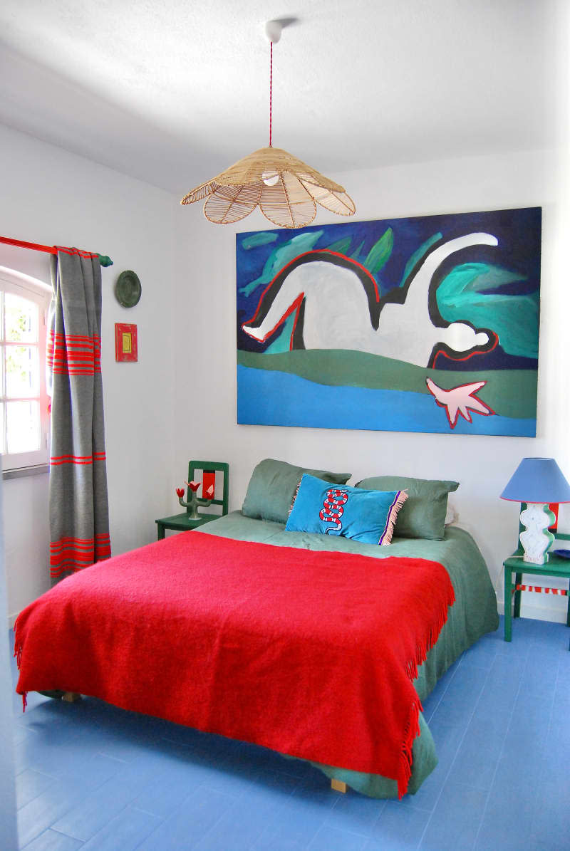Artwork and rattan lamp hung above bed with green and red bedding in white bedroom with blue floor.
